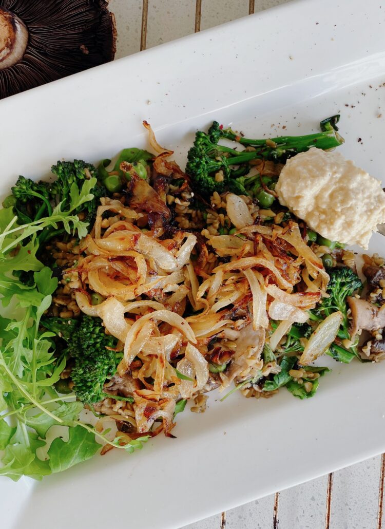 A nutritious plate of freekeh cooked with green peas and mushrooms, topped with fresh rocket, caramelized onions, hummus, and a sprinkle of thyme.