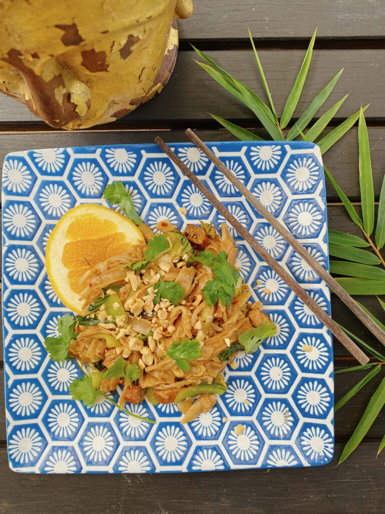 A plate with pad see ew, famous thai dish on it, featuring peanuts, cilantro, noodles and tofu on a brown background.