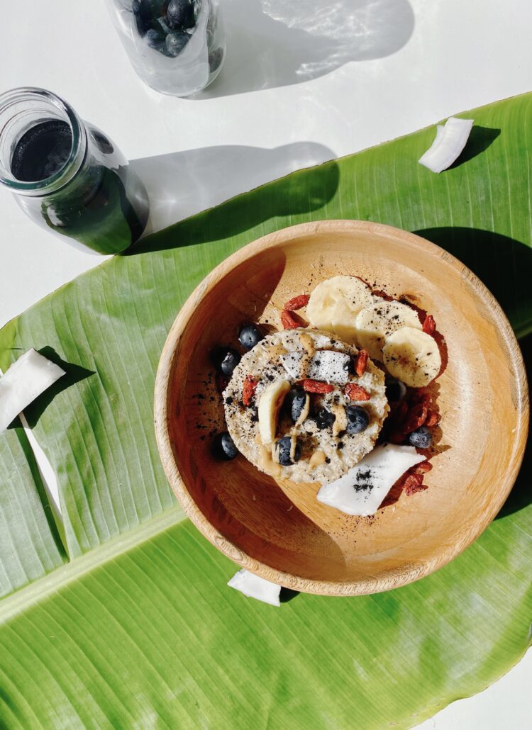 A bowl of mixed berries (blueberries and goji berries), shredded coconut, and oats served on a banana leaf, accompanied by pieces of charcoal on the side.