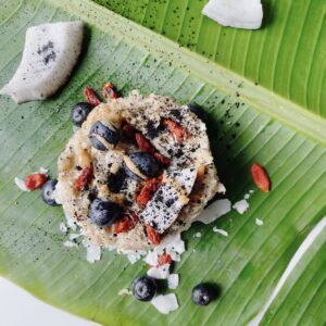 A bowl of vegan oats topped with goji berries, blueberries, and coconut, served on a vibrant green banana leaf with shredded coconut and sprinkled with activated charcoal.