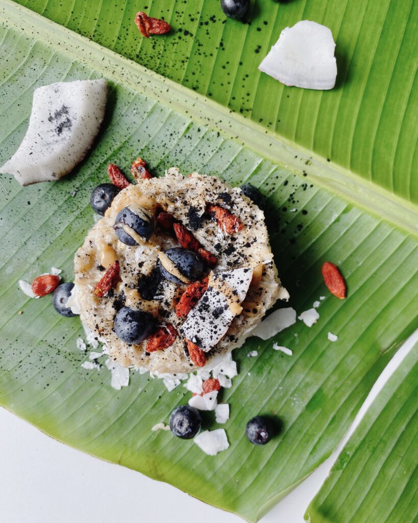 A high protein bowl of oats topped with goji berries, blueberries, and coconut, served on a vibrant green banana leaf with shredded coconut and sprinkled with activated charcoal.