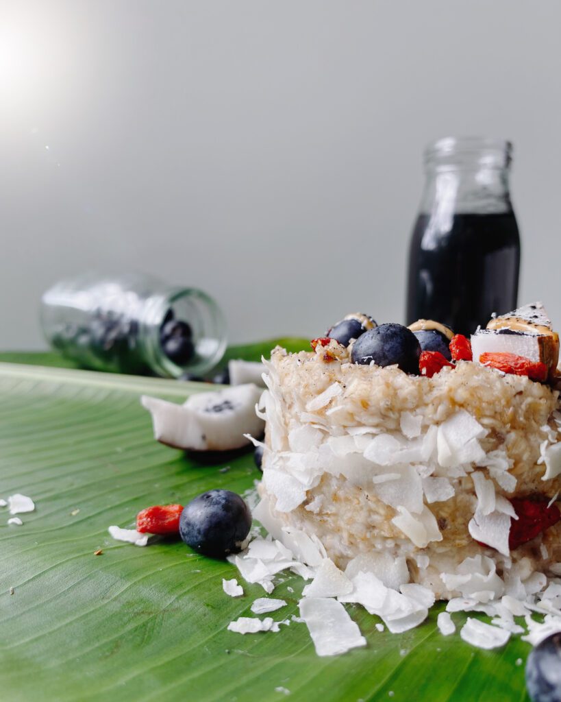 Vegan oats topped with a colorful assortment of fresh berries, shredded coconut, and activated charcoal powder, presented on a tropical banana leaf.