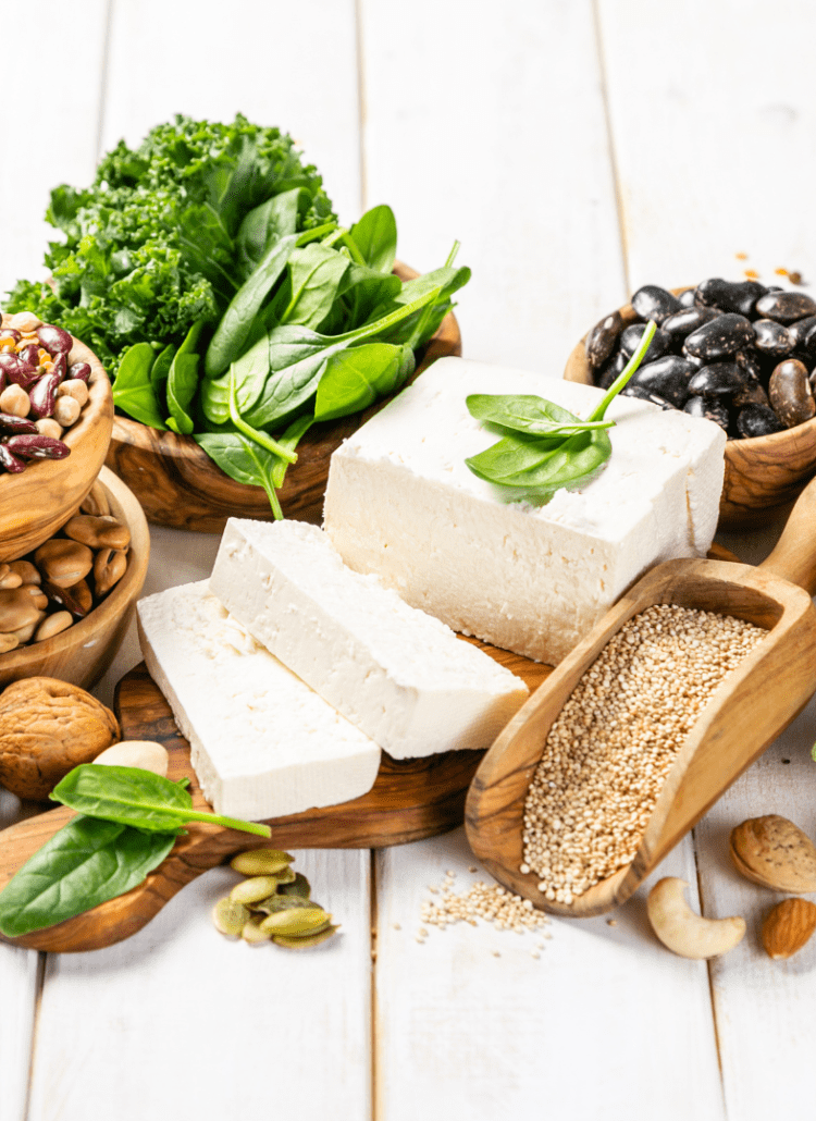 A selection of plant-based protein sources displayed on a wooden table. There are bowls containing various legumes, nuts, and seeds, along with fresh greens like kale and spinach. A block of tofu is also prominently featured.