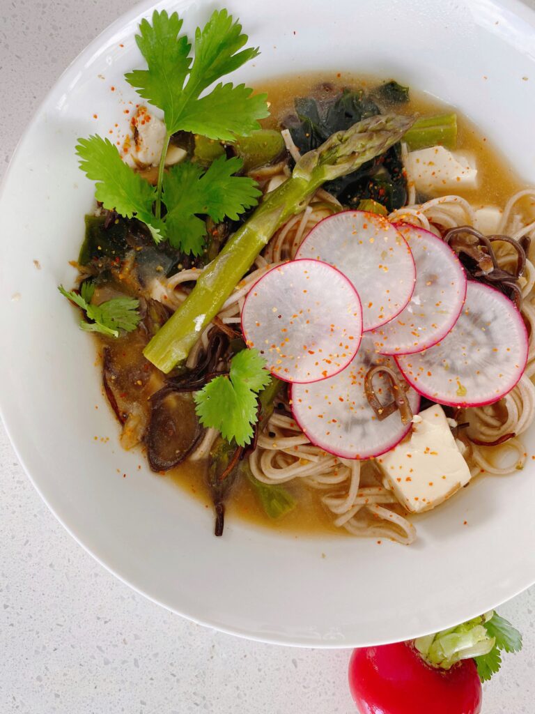 A bowl of noodle soup, containing thin, white noodles and is garnished with thinly sliced radishes, chunks of tofu, and a few leaves of fresh cilantro. There are also mushrooms, asparagus stalks, and dark green leafy vegetables visible in the mix. The bowl is placed on a light grey table, and there's a radish on the table.