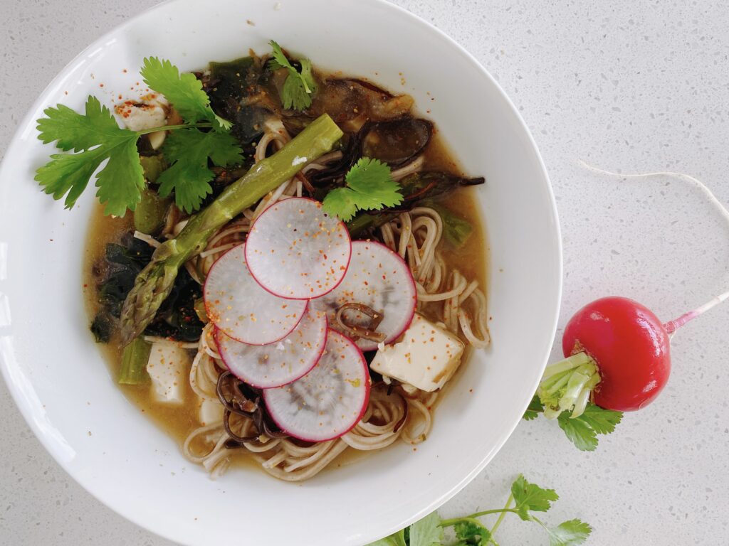 A top-down view of a bowl of noodle soup placed on a light grey countertop. The soup contains a variety of ingredients including soba noodles, sliced radishes, blocks of tofu, and greens, garnished with cilantro leaves. A single asparagus stalk rests a top the soup. Specks of red seasoning sprinkle the surface of the broth, adding a touch of color.