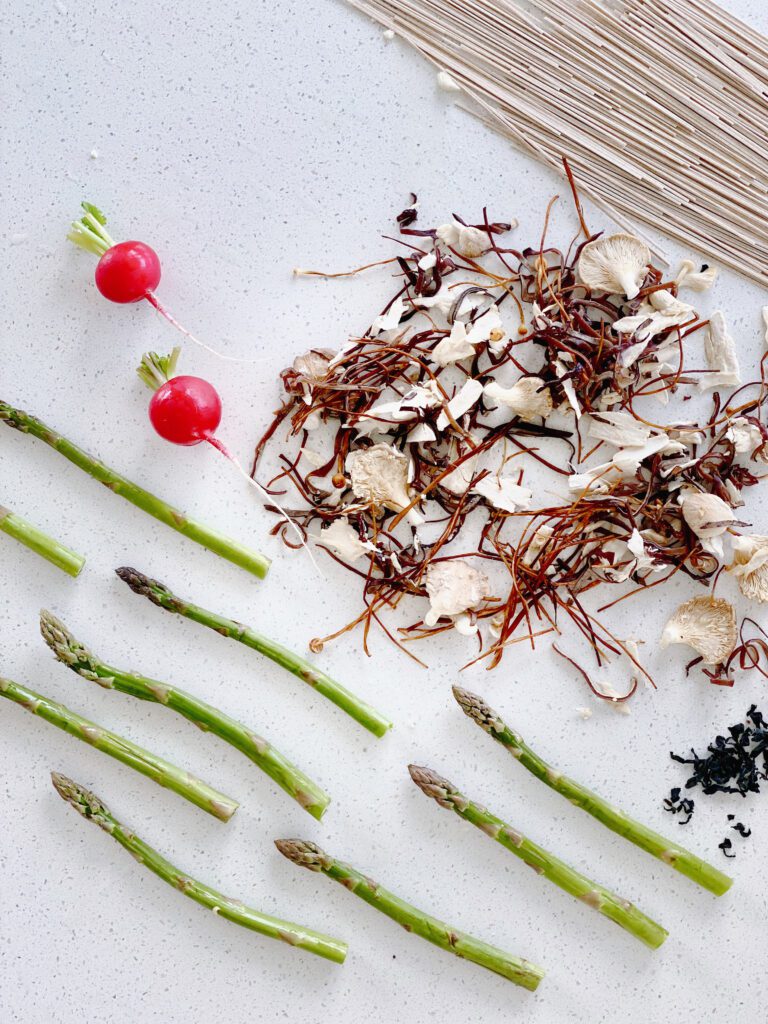 A flat lay of various raw ingredients artfully arranged on a light grey textured surface. There are several bright green asparagus stalks laid out in parallel lines with their tips pointing in various directions. To the left, two vibrant red radishes with green stems are placed, one with its root elegantly curved. In the center, there's a heap of chopped mushrooms intermingled with thin red chili. Near the top right corner, a bundle of uncooked soba noodles is neatly aligned.