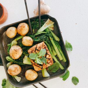 A plant-based meal, prominently featuring a golden-browned block of tofu that has been expertly pan-seared to create a delicious crust. It's garnished with green herbs like baby spinach leaves, adding a fresh, vibrant contrast. Accompanying the tofu are several round, lightly-browned potatoes and an array of green baby bok choy. The food is arranged in a non-stick skillet, hinting that it might be freshly prepared and ready to be served.