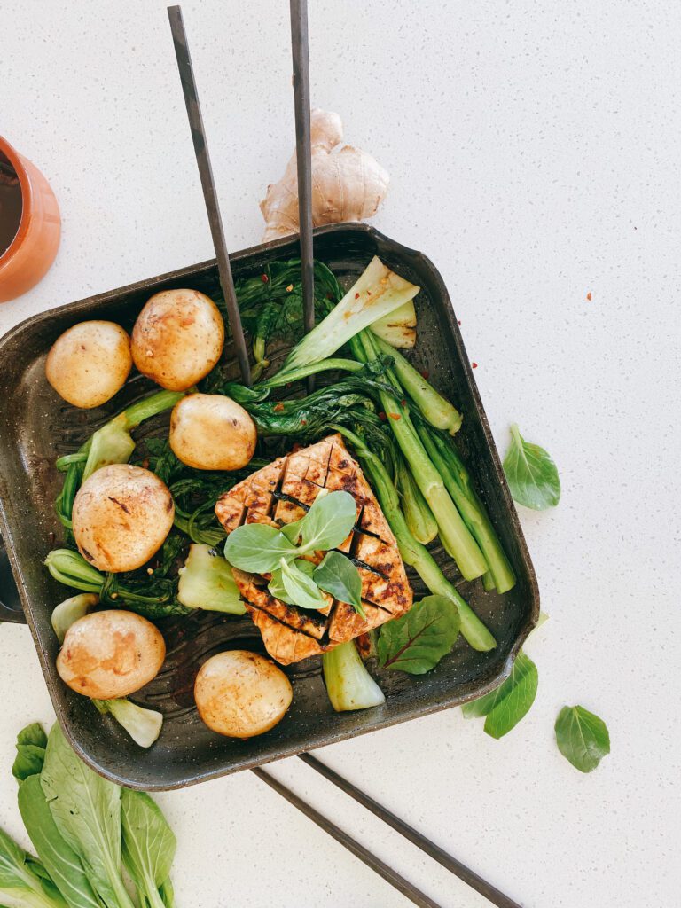 A plant-based meal, prominently featuring a golden-browned block of tofu that has been expertly pan-seared to create a delicious crust. It's garnished with green herbs like baby spinach leaves, adding a fresh, vibrant contrast. Accompanying the tofu are several round, lightly-browned potatoes and an array of green baby bok choy. The food is arranged in a non-stick skillet, hinting that it might be freshly prepared and ready to be served.