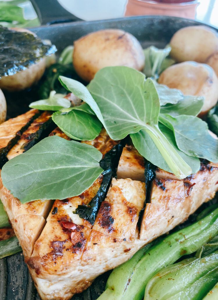 This is a vibrant and appetizing image of a pan-seared meal consisting of a grilled tofu steaks topped with fresh herbs, surrounded by a selection of vegetables. There are whole, golden-browned potatoes and green baby bok choy.