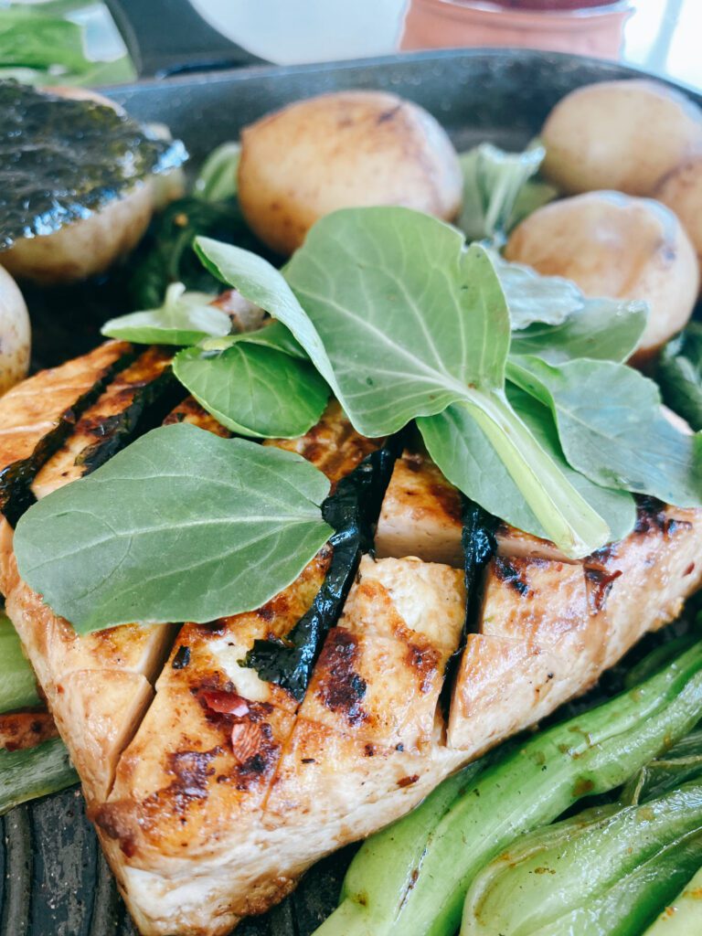 This is a vibrant and appetizing image of a pan-seared meal consisting of a grilled tofu steaks topped with fresh herbs, surrounded by a selection of vegetables. There are whole, golden-browned potatoes and green baby bok choy.