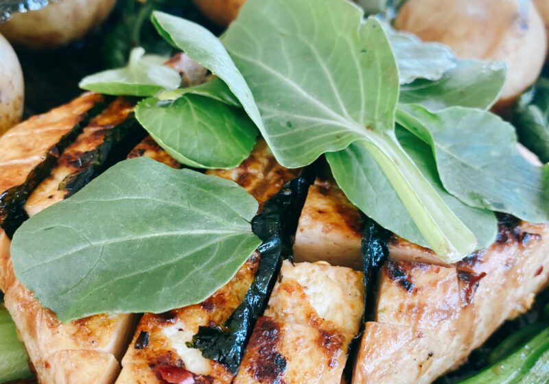 This is a vibrant and appetizing image of a pan-seared meal consisting of a grilled tofu steaks topped with fresh herbs, surrounded by a selection of vegetables. There are whole, golden-browned potatoes and green baby bok choy.