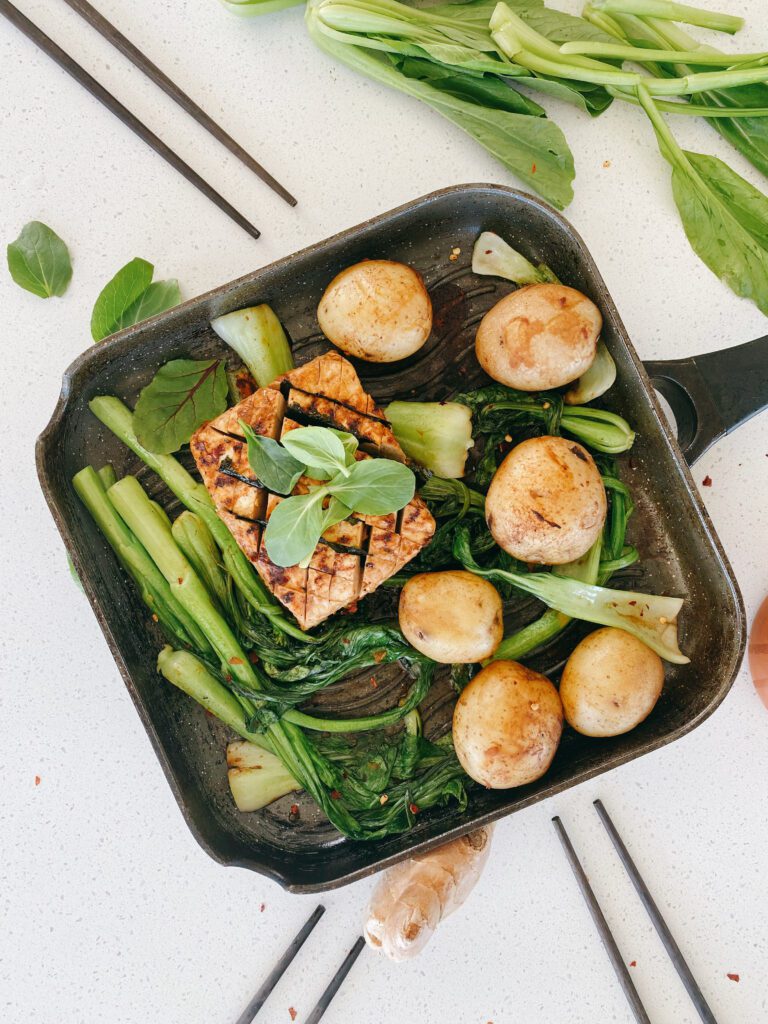 A healthy and appealing vegan dish consisting of a pan-seared block of tofu with a golden crust, accompanied by round, roasted potatoes and a side of green bok choy. The tofu has clear grill lines, suggesting it was cooked to achieve a caramelized exterior, while the potatoes show a light browning indicative of roasting or pan-frying. The bok choy maintains its vibrant green color, with slight charring on the edges, pointing to a brief cooking time to preserve its texture and color. This dish is presented in a modern, non-stick skillet, which underscores its readiness to be served. 